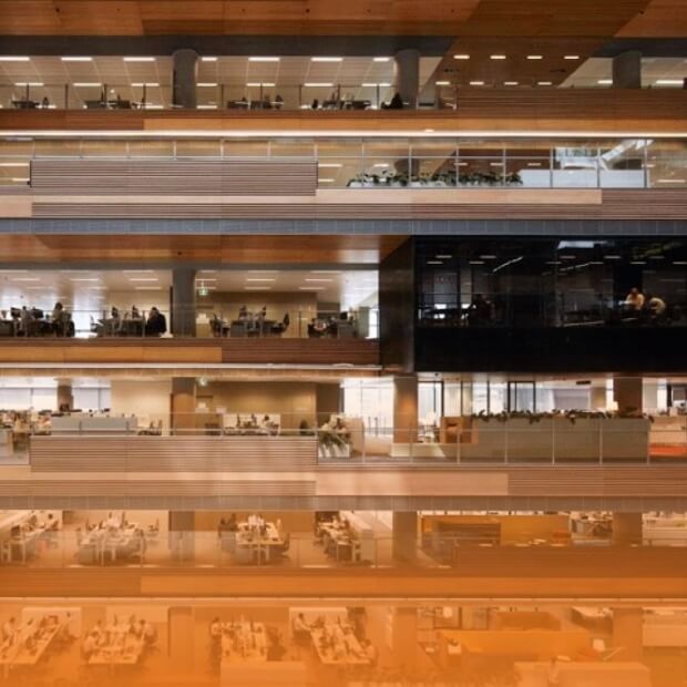 A multi-level modern office building with glass walls reveals a nurturing culture of innovation. The exterior showcases several floors, with illuminated interiors where plants and office equipment are visible through large windows, highlighting people creatively working at their desks.