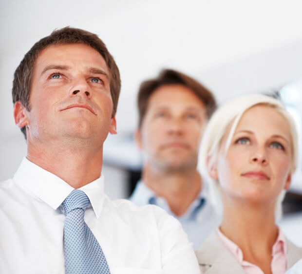 Three people in business attire, embodying an upskilling mindset, look confident and focused as they gaze upwards. The image captures a man in a white shirt and tie sharply in the foreground, with a man and woman slightly blurred behind him, set against an out-of-focus background.