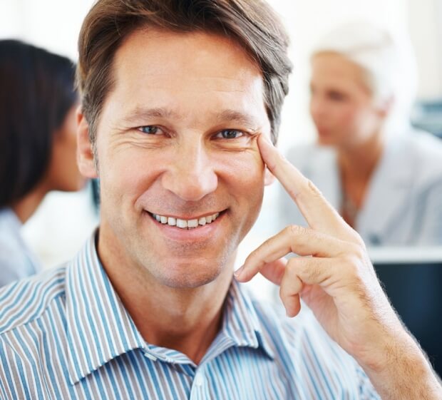 A smiling man in a striped shirt, exuding emotional intelligence, rests his finger on his cheek as he sits in an office environment with two blurred colleagues in the background.