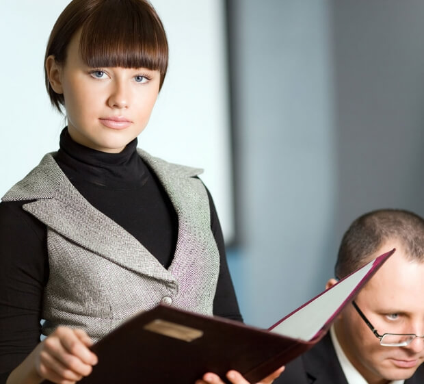 A woman with short brown hair, dressed in a black turtleneck and gray vest, is confidently holding an open folder. She stands beside a man with glasses who is looking down, as if engaged in informal coaching. The background is blurred.