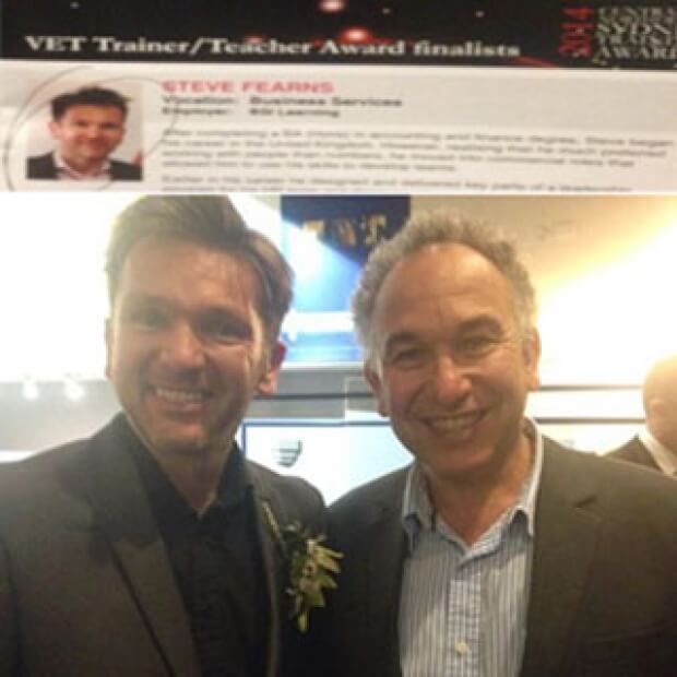 Two smiling men pose together indoors, exuding a sense of leadership. One man is wearing a flower on his jacket, and a poster titled VET Trainer/Teacher Award finalists is partially visible above them.