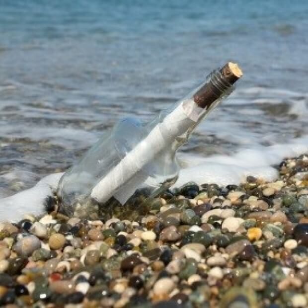 A glass bottle containing a rolled-up message rests on a pebble-covered shoreline. Demonstrating clear communication, the scene captures how waves gently lap around the bottle, while the ocean stretches out in the background, inviting us to imagine messages that aim for clarity and purpose.